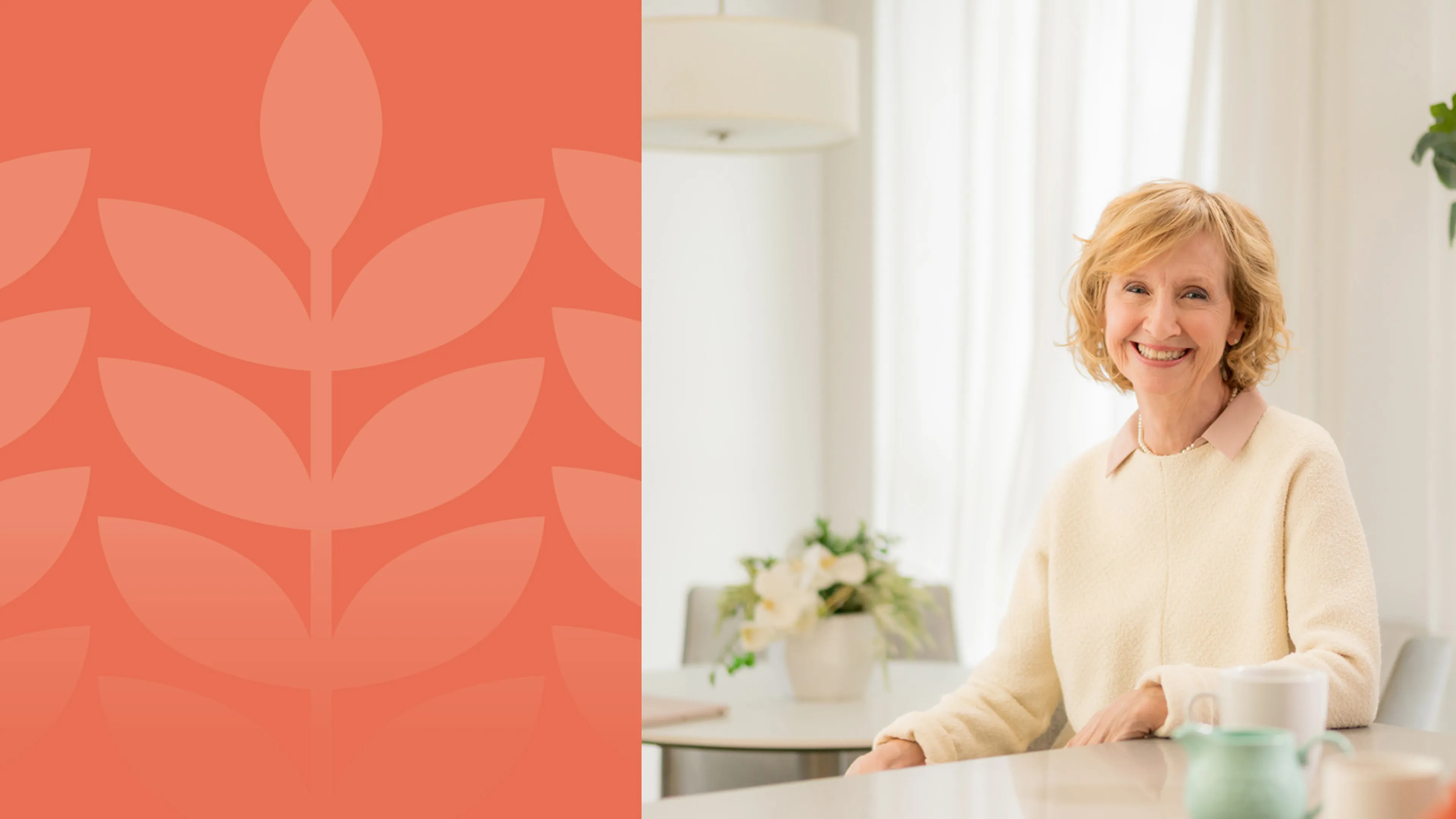 An older lady is sitting at a white kitchen table, smiling at the camera.