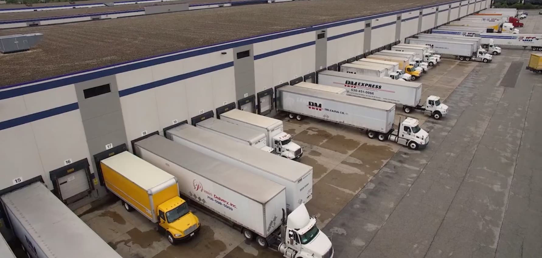 An aerial photo of a major distribution center showing a long line of white semi-trucks and box trucks at the loading dock, illustrating a large-scale trucking operation.