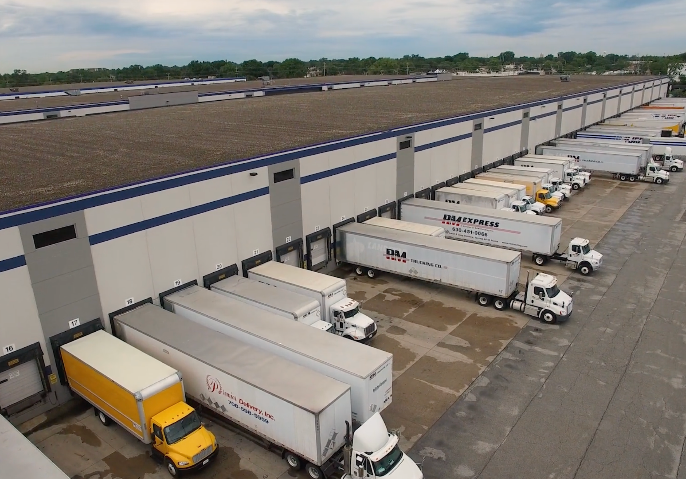 An aerial photo of a major distribution center showing a long line of white semi-trucks and box trucks at the loading dock, illustrating a large-scale trucking operation.