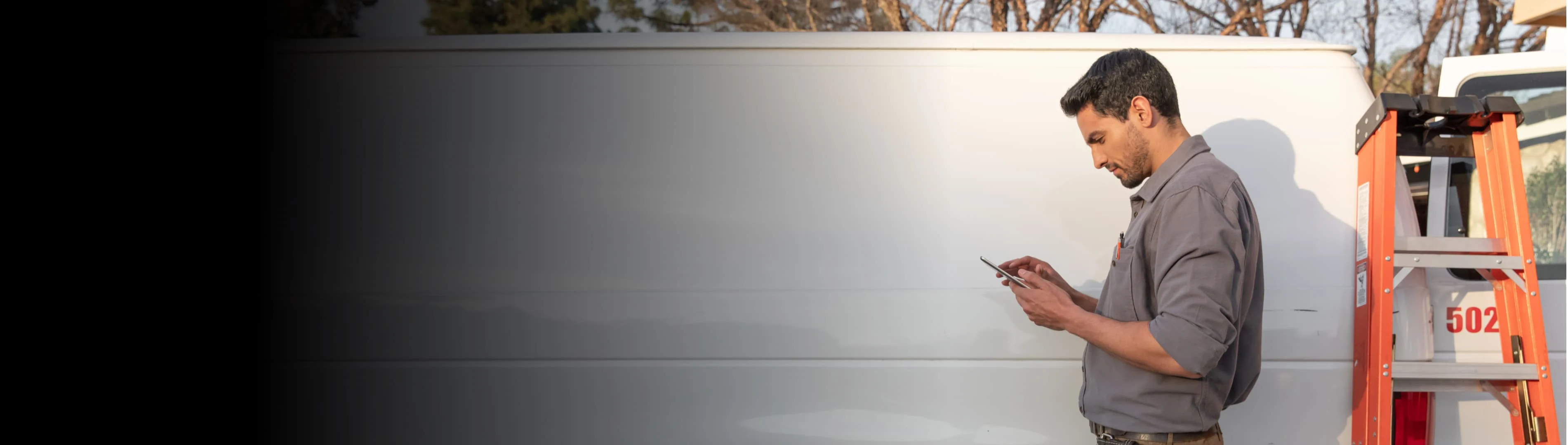 Service worker in a gray shirt standing next to a white van, looking at a phone or tablet.