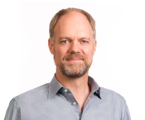 Headshot of a smiling man, Peter Mitchell, with a light beard, wearing a light blue collared shirt.