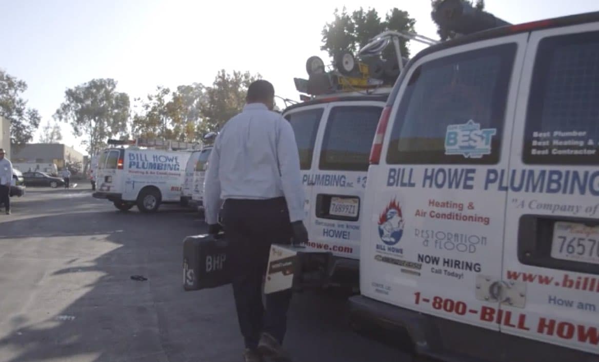 A male service technician carrying a toolbox walks past a row of white service vans, each branded with "Bill Howe Plumbing, Heating & Air Conditioning."