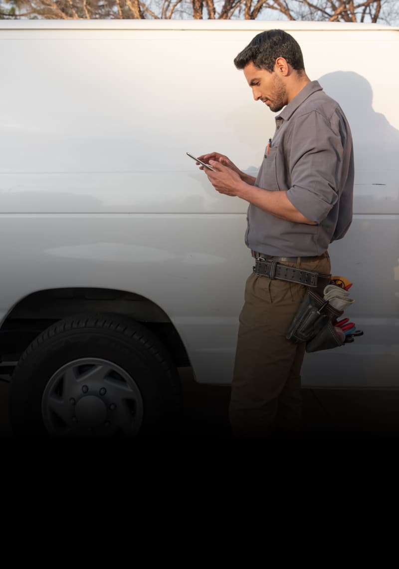 Service worker in a gray shirt standing next to a white van, looking at a phone or tablet.
