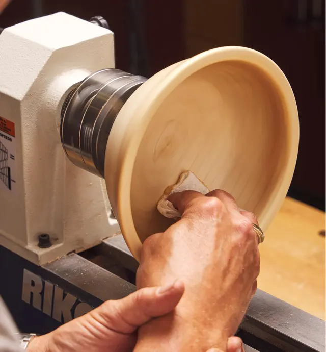 A woodturner uses Yorkshire Grit abrasive paste on a paper towel to polish a wooden bowl on a lathe