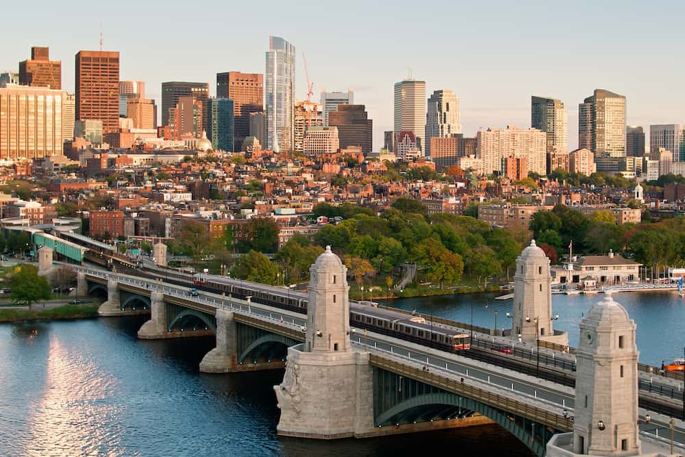 A wide cityscape showing a bridge over a river with a train crossing it in the foreground and a skyline of tall buildings and residential houses behind, illuminated by warm evening light.
