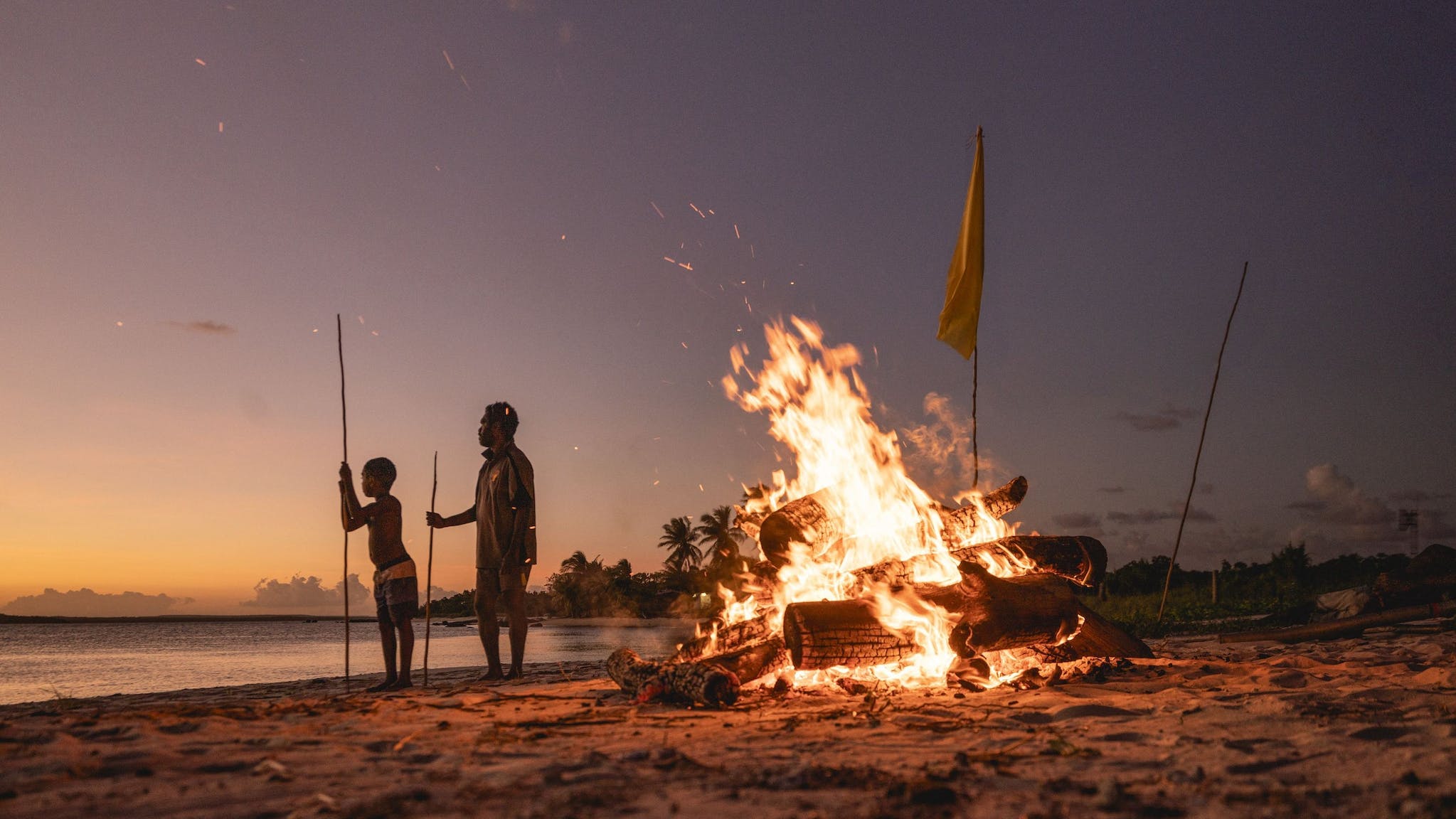 Aboriginal man and child holding spears next to a campfire on the beach in East Arnhem Land