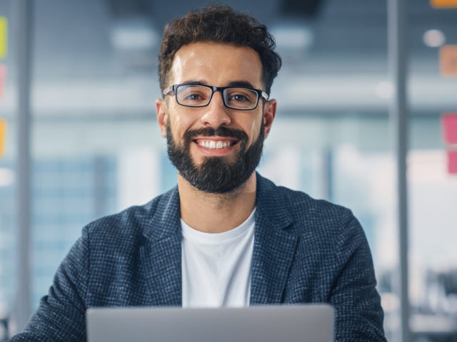 Hombre con gafas sonriendo frente a una laptop en una oficina.