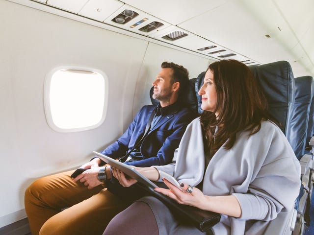 A young couple is sitting on a plane and looking out the window with anticipation, holding their moving abroad checklist.