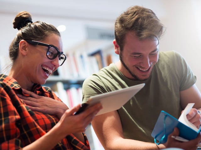 A young female adult and a male friend in casual clothing are laughing and learning how to become a polyglot, together.