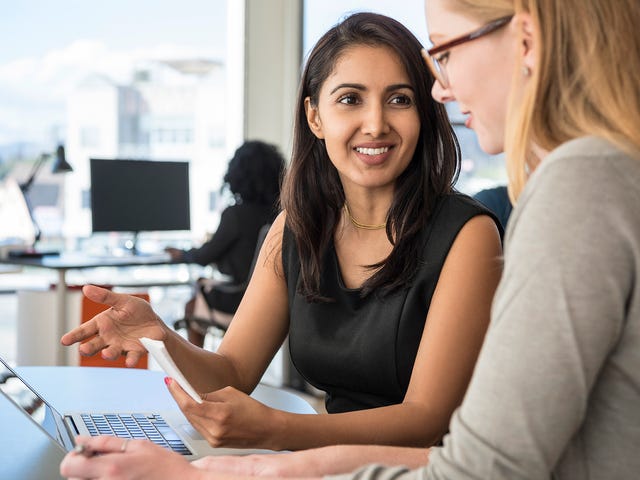 Two ladies of different ethnic backgrounds smiling and chatting over a laptop in an office space.