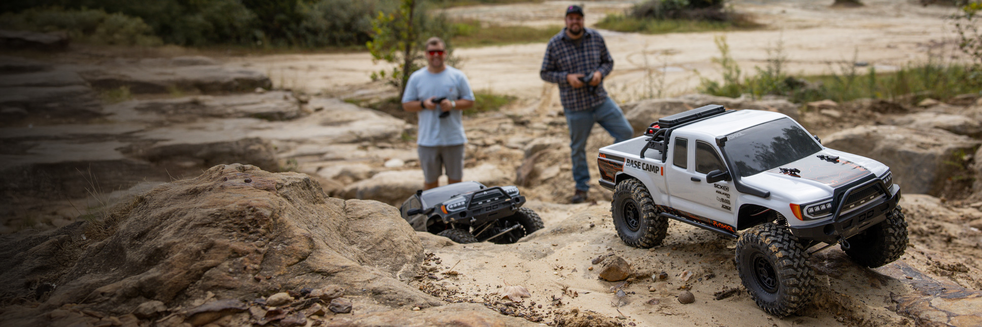 Action shot of the Axial 1/10 SCX10 III Base Camp 4X4 Ready-To-Run Brushed Rock Crawlers in White and Gray climbing over rocks with two people standing in the background holding transmitters in their hands Action shot of the Axial 1/10 SCX10 III Base Camp 4X4 Ready-To-Run Brushed Rock Crawlers in White and Gray climbing over rocks with two people standing in the background holding transmitters in their hands