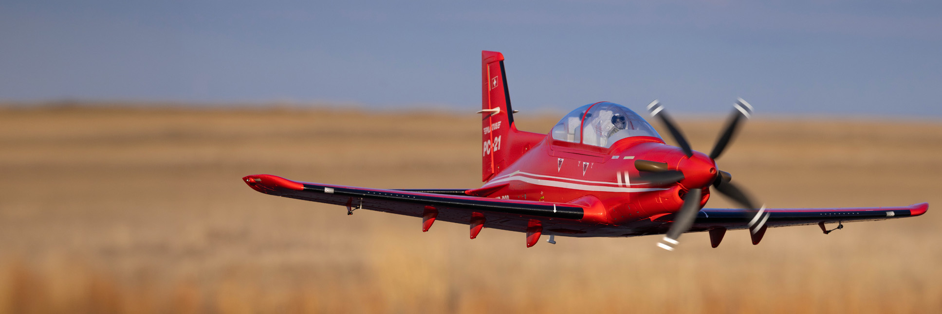 Action shot of the E-flite Pilatus PC-21 1.3m flying above a tarmac next to a field Action shot of the E-flite Pilatus PC-21 1.3m flying above a tarmac next to a field