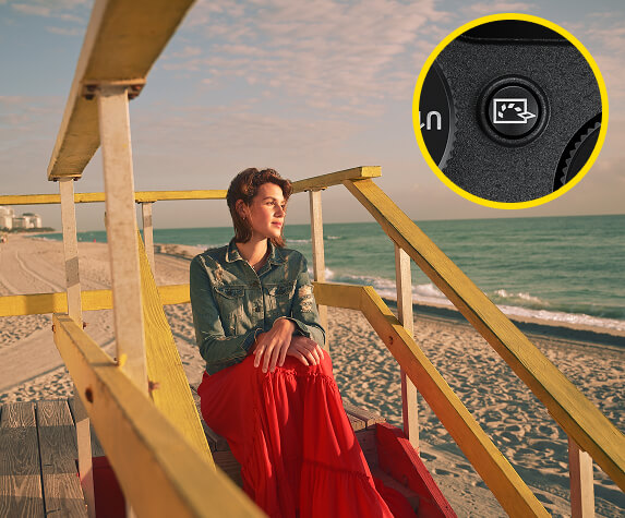 A woman in a long red dress sits on yellow-painted wooden steps on a sandy beach, looking out to the ocean.