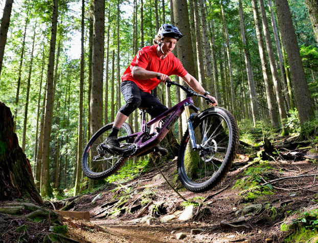A male mountain biker is shown mid-air while riding through a lush forest trail.