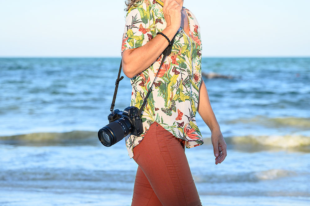 photo of a woman holding a camera over her shoulder walking on the beach
