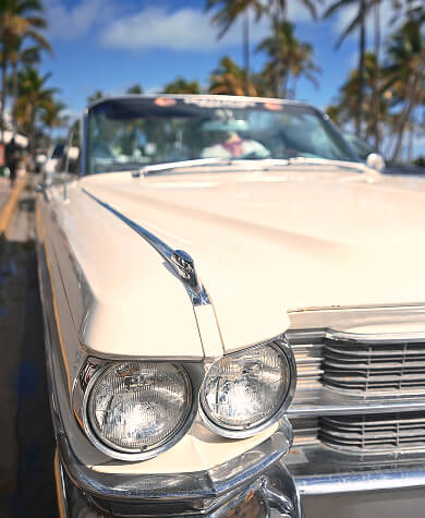 A cream-colored vintage car sits on a Miami street with blue skies and palm trees in the background.