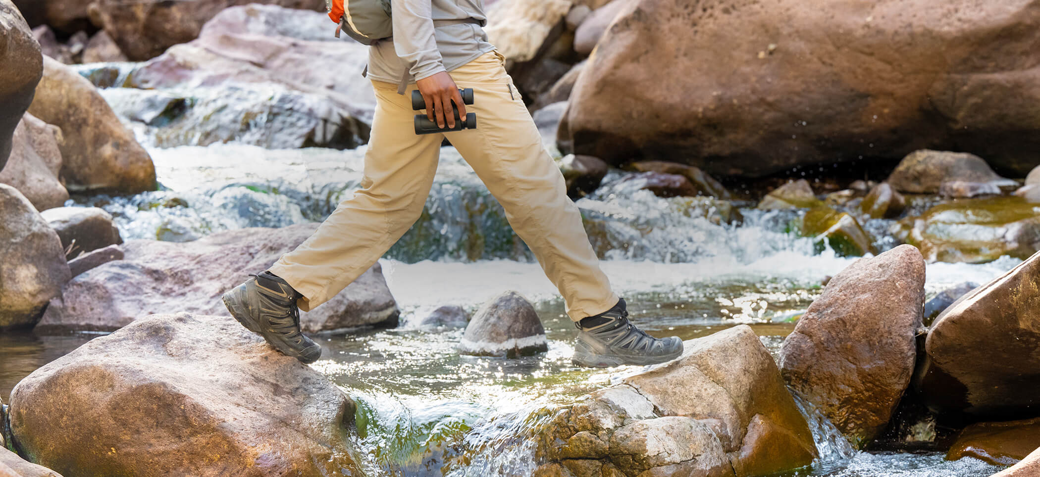 Photo of a man walking over water and rocks, holding a pair of PROSTAFF P3 8X42 binoculars