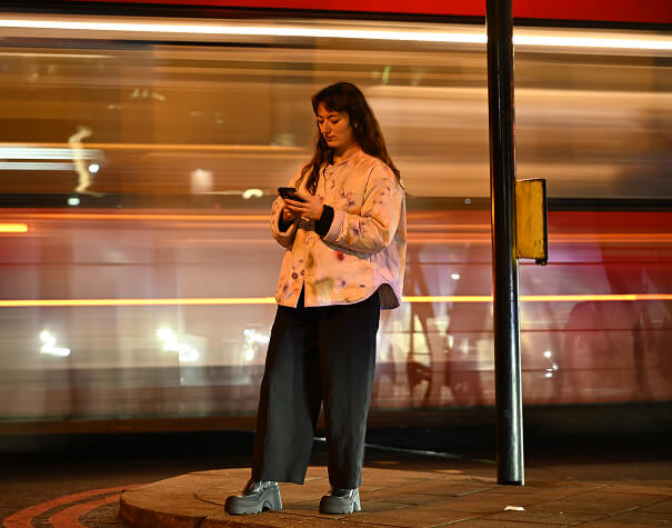 A young woman looking at a smartphone on a city walkway while a bus passes behind her.