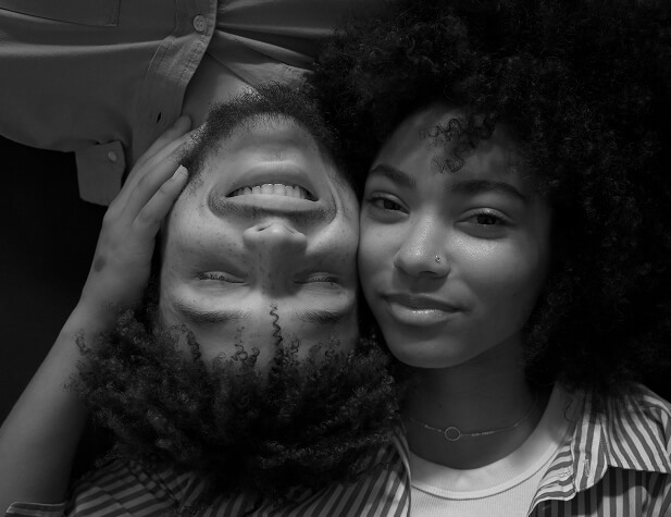A black and white photo of a young couple lying on their backs with faces gently touching.