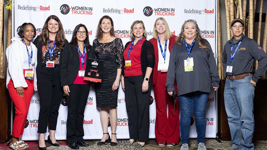A group of eight women stands in front of a banner that reads "Women in Trucking" and "Redefining the Road." 