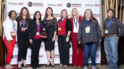 A group of eight women stands in front of a banner that reads "Women in Trucking" and "Redefining the Road."