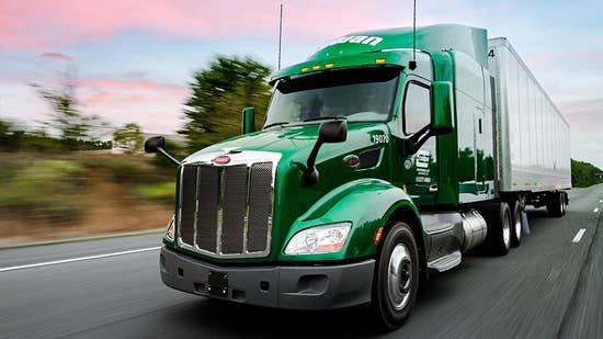 cowan anniversary 2025_hero.jpg A green Cowan truck and trailer drive down the highway at sunrise, with pink hues of color in the sky behind it