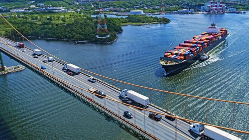 An aerial view of a large cargo ship carrying numerous colorful shipping containers sailing under a suspension bridge. The bridge has multiple lanes with various vehicles, including semi trucks and cars, traveling across it.