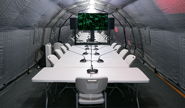 Interior of a portable military briefing tent with a conference table, microphones, and a screen displaying a tactical map