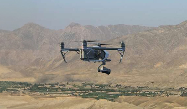 A military quadcopter drone with a camera payload flying over an arid, mountainous landscape