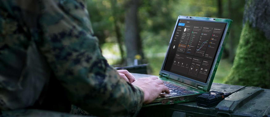 A soldier in camouflage uniform using a ruggedized laptop outdoors in a wooded environment