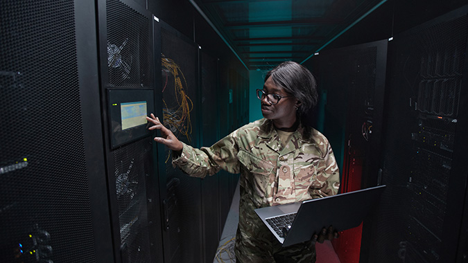A soldier in camouflage uniform holds a laptop while interacting with a server rack panel in a dark data center.
