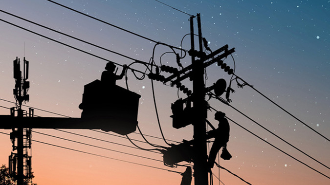 Silhouettes of two utility workers servicing a power pole at dusk, one in a bucket lift and one climbing the pole.