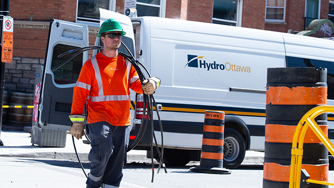 A Hydro Ottawa utility worker in an orange safety vest and green hard hat carries electrical cables on a city street.