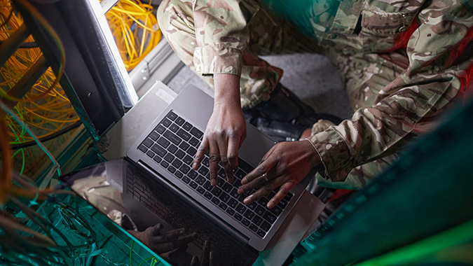 Overhead view of a soldier in camouflage typing on a laptop surrounded by network cables