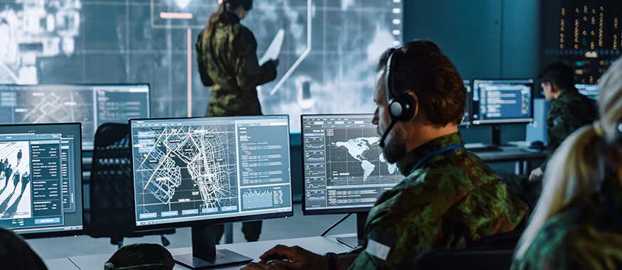 Military personnel in a darkened operations center monitoring multiple screens displaying maps and surveillance data