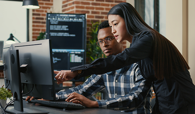 Two IT professionals collaborating at a computer workstation to configure and monitor secure network defense systems