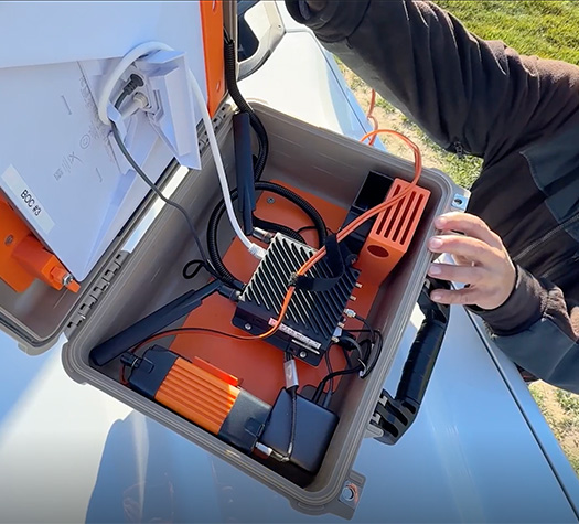 An Idaho Power technician inspects an open outdoor equipment case containing a router and cables in the field.