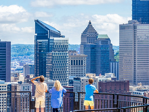 Drie mensen staan op een uitkijkpunt en kijken uit over het panorama van de stad Pittsburgh 