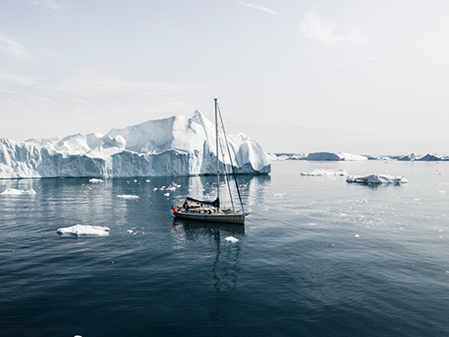 A sailboat is making its way across a body of water in Greenland, with icebergs to the background of the picture 