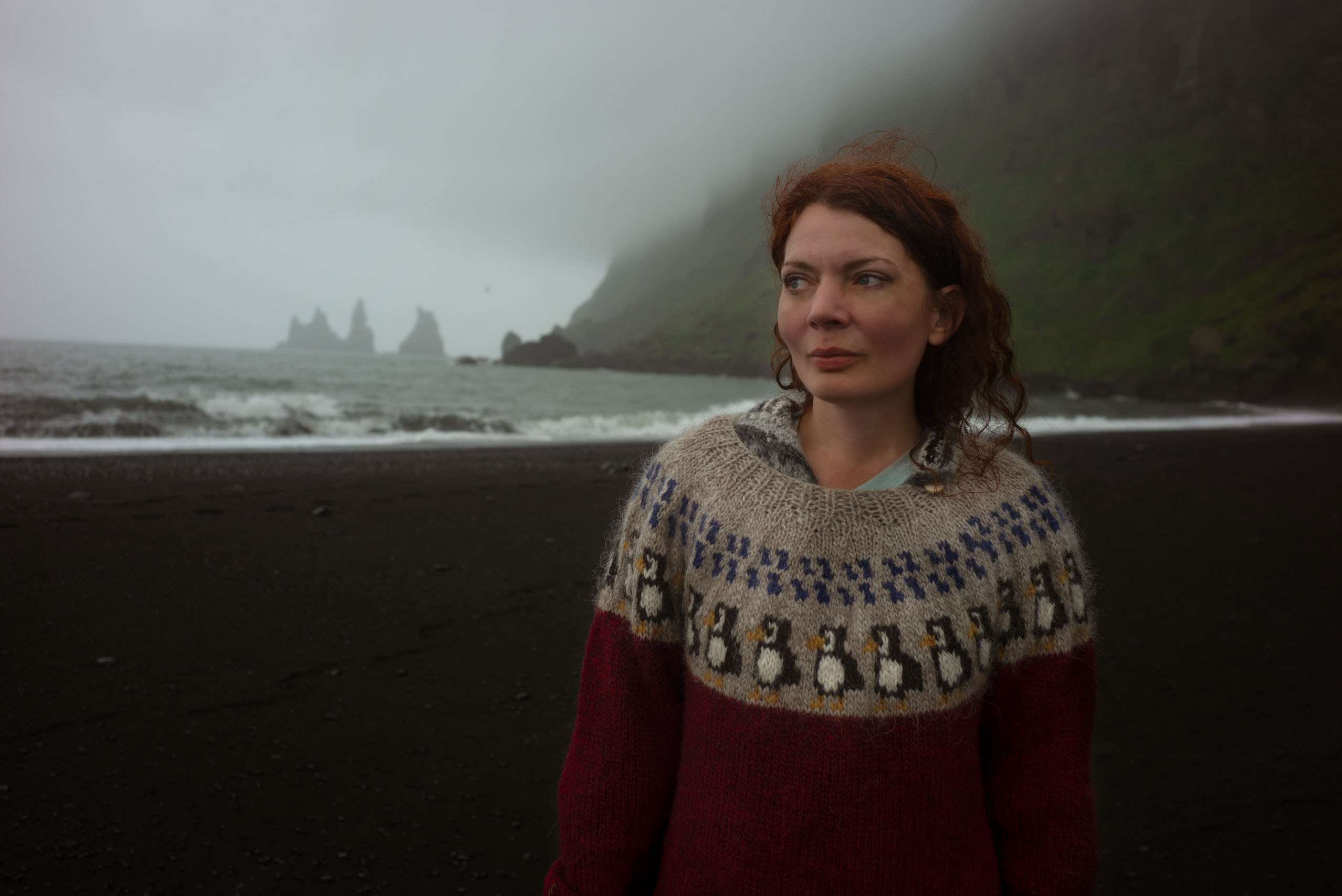 A woman wears a lopapeysa with puffin pattern, standing on a black beach at Vík.