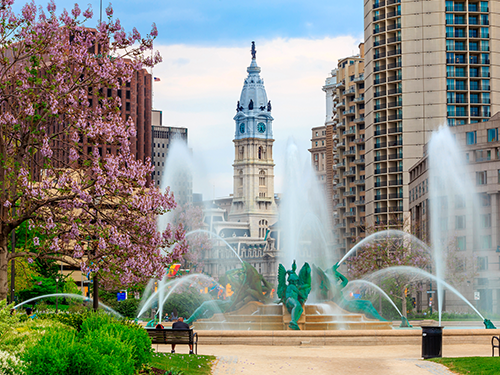 A scene of Philadelphia in the summer, with lush greenery and pink flowers in frame, set against the historic buildings 