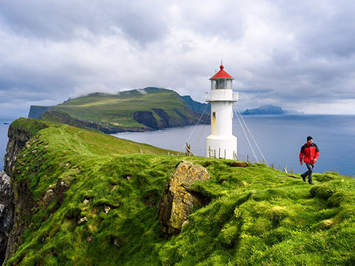 Un homme en veste de pluie rouge traverse une crique herbeuse d’où l'on aperçoit au loin un phare blanc et rouge 