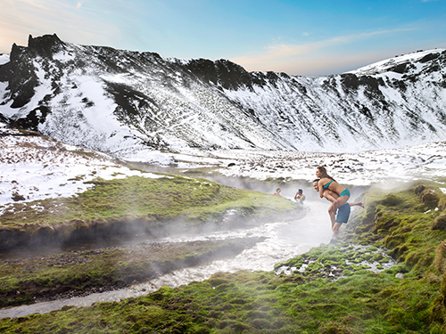 Reykjadalur geothermal valley in Iceland, here pictured with people bathing in the water and a couple to the right of the picture with a female getting a kiddy-back from her male companion 