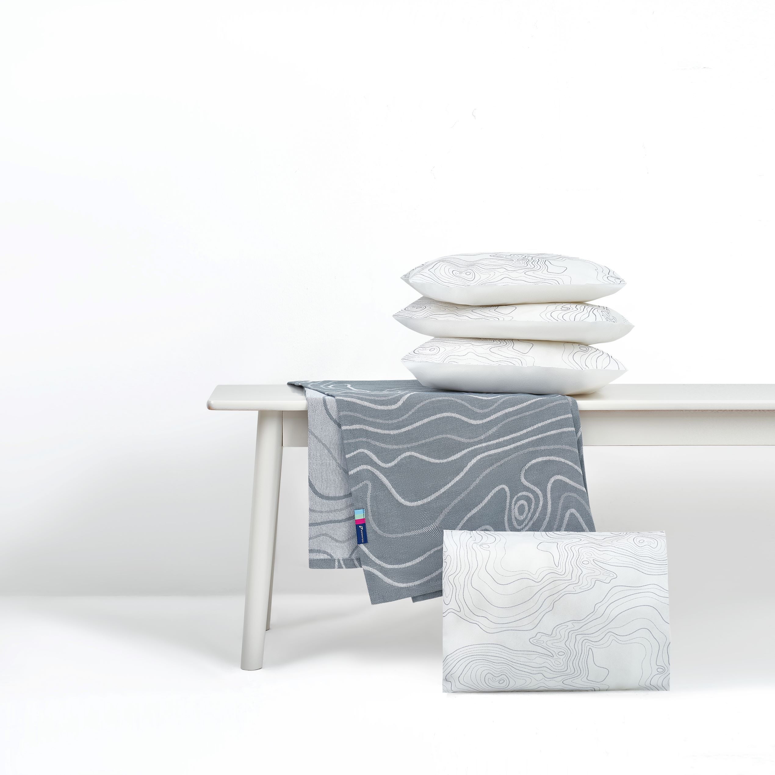 Our new onboard Economy textiles set shows a gray blanket with white topographic lines, and white pillows with faint grey topographic lines, photographed on a white bench