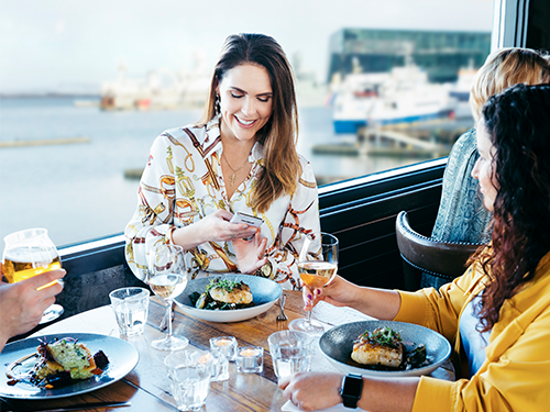 A woman dines with friends, eating fish by the harbour in Reykjavík with Harpa in the background