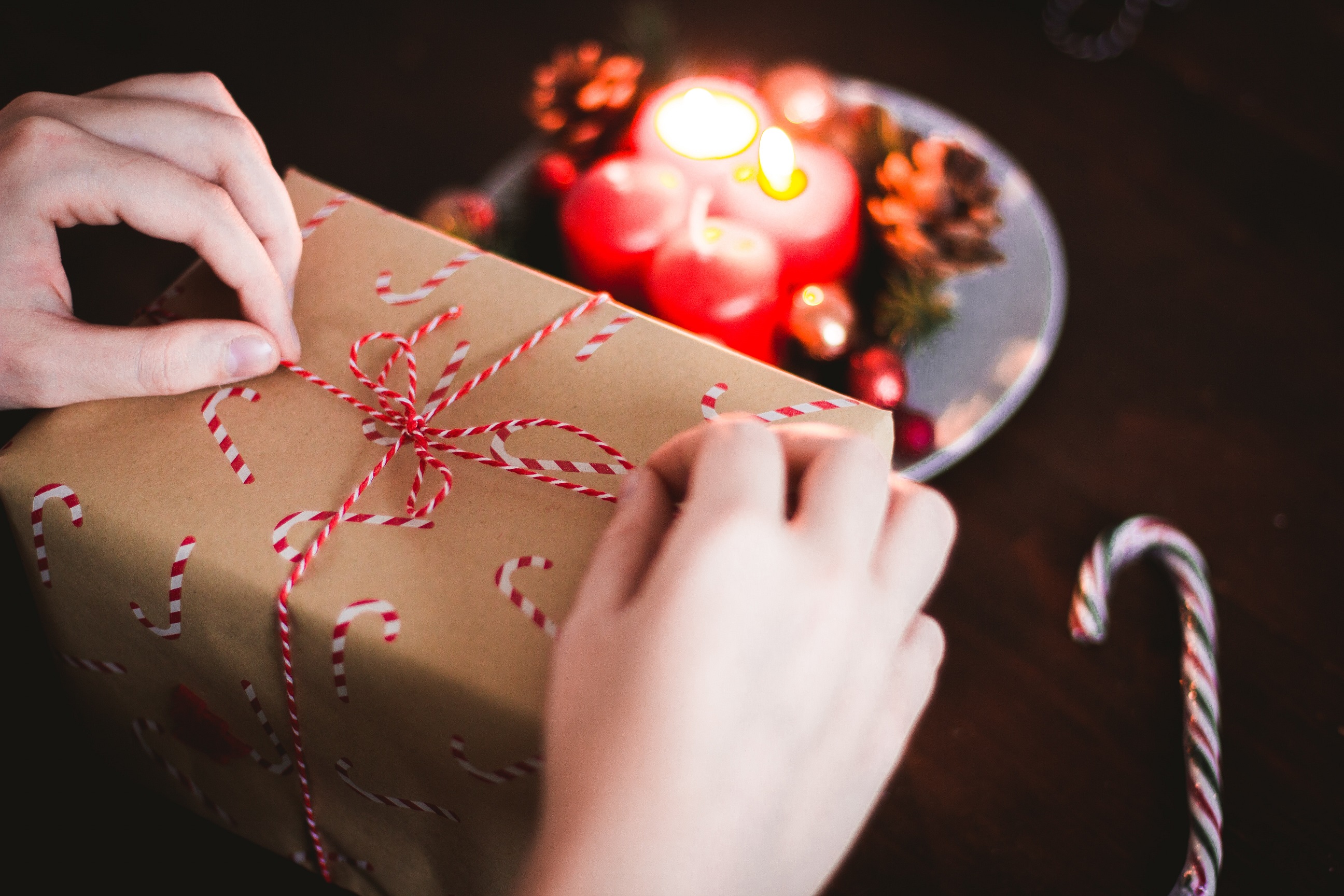 Two white hands pictured unfastening the red ribbon bow on the top of a brown paper packaging parcel which has red and white candy canes decorating it
