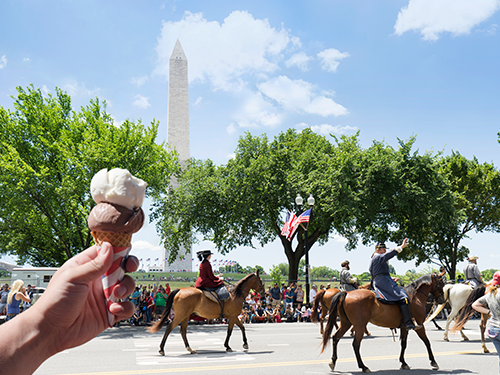 A hand holds a chocolate and vanilla ice cream up in front of a scene of several people on horses