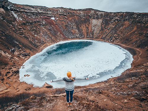 Kerið Crater on the Golden Circle route, here pictured frozen over. with a girl with her back to us wearing a yellow hat and holding her arms outstretched 