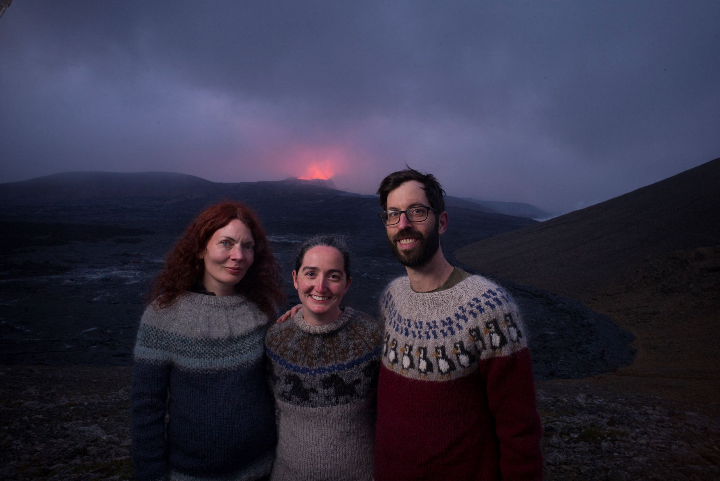 Three people - a man and two women - are all wearing handkniited sweaters, and stand in the foreground while in the distance the Fagradallsfjall volcano is erupting