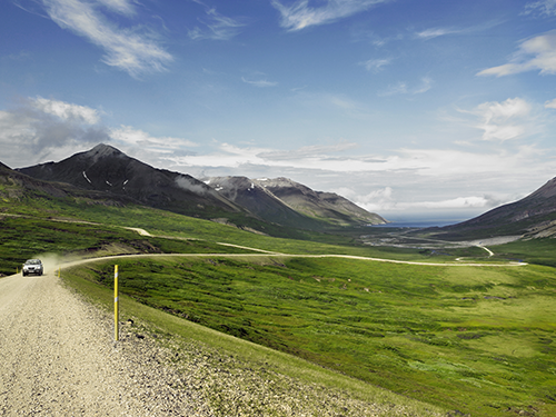 A car is driving through the Eastfjords towards the camera, with fjords pictured in the background  